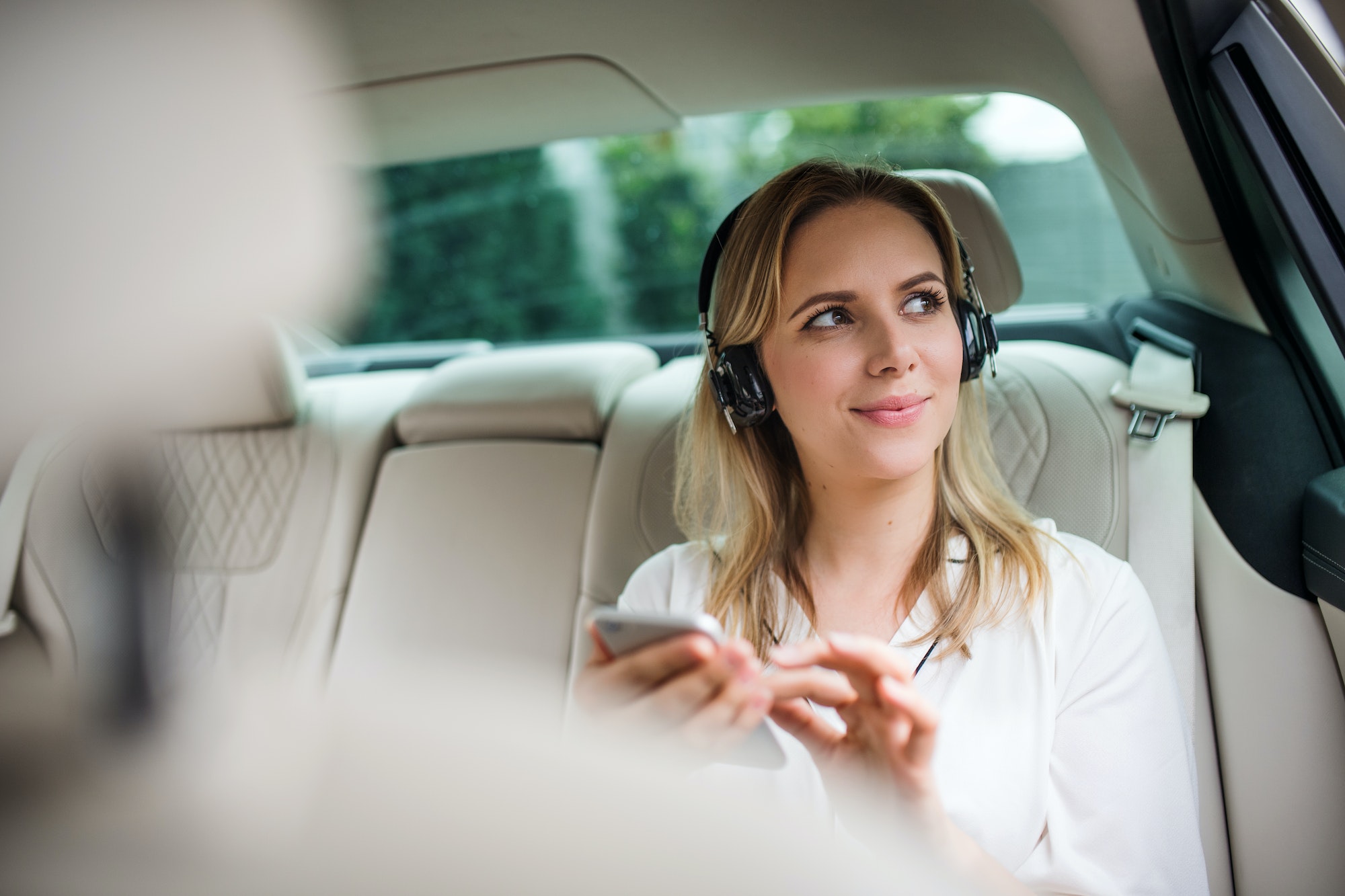 Business woman with smartphone and headphones sitting on back seats in taxi car.